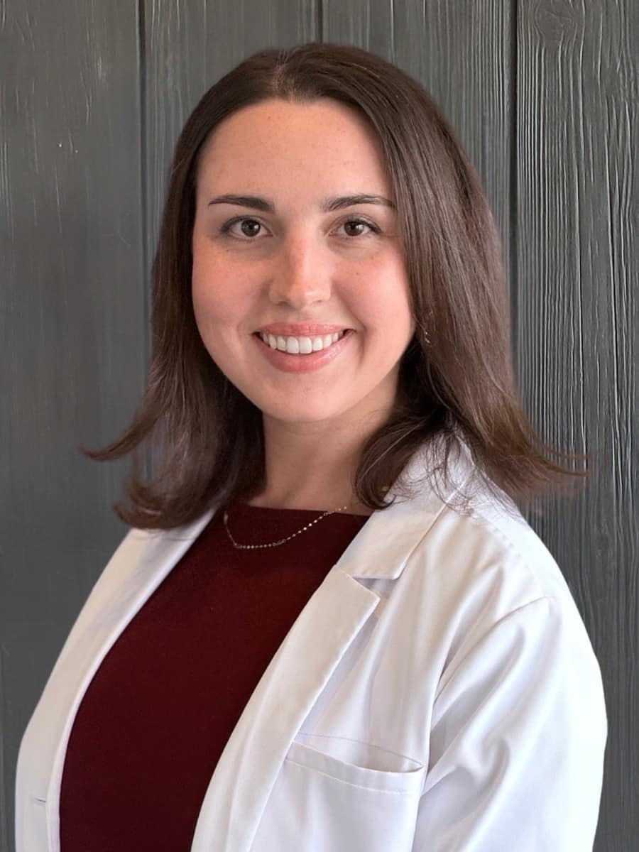 Vital-care-samantha-scalia Smiling female healthcare professional wearing a white coat in front of a gray wooden background.